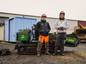 Two workers are standing beside a stump grinder machine, both wearing protective gear. This scene illustrates the process of how to grind a tree stump.