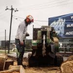 Urban tree lopper operating stump removal solution with heavy machinery near workshop.