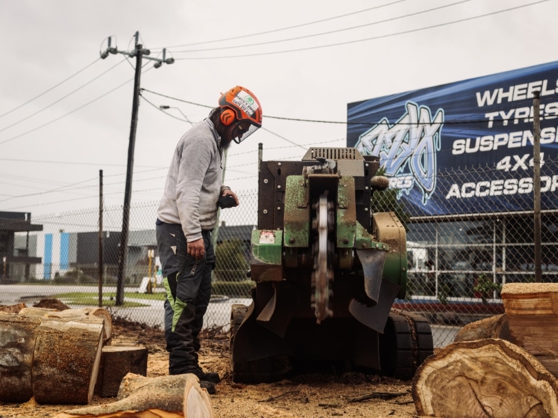 Urban tree lopper operating stump removal solution with heavy machinery near workshop.