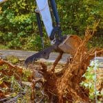Excavator claw performing tree root removal on a rural property with red soil and forest background